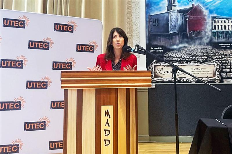 National Institute of Justice Director Nancy La Vigne speaks from behind podium. UTEC Inc banner in background.