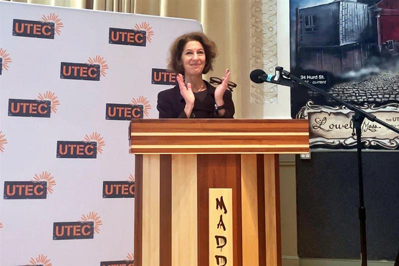 Assistant Attorney General Amy L. Solomon applauds from behind podium. UTEC Inc banner in background.