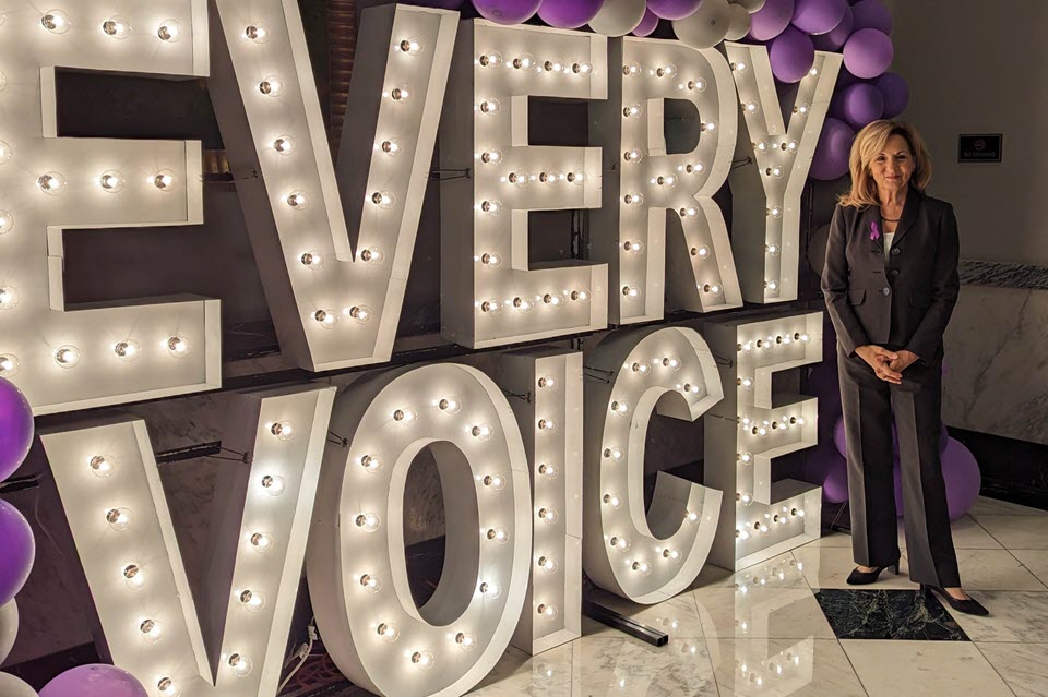 OVC Director Kris Rose stands in front of large lighted sign that reads Every Voice