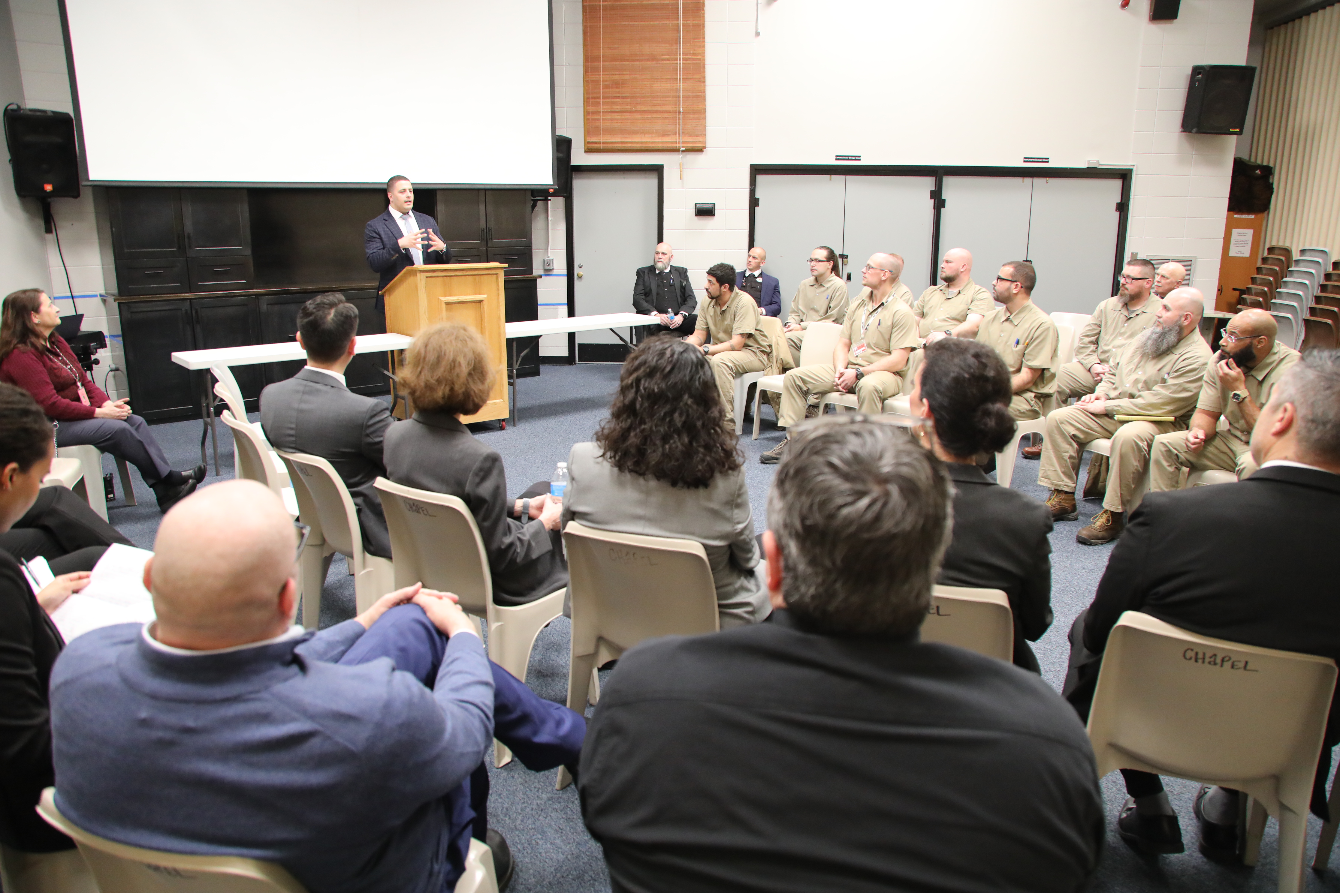 A speaker stands at a podium addressing a mixed group of inmates and officials seated in a large room during a correctional education event. The audience includes both uniformed inmates and formally dressed officials, attentively listening to the presentation.