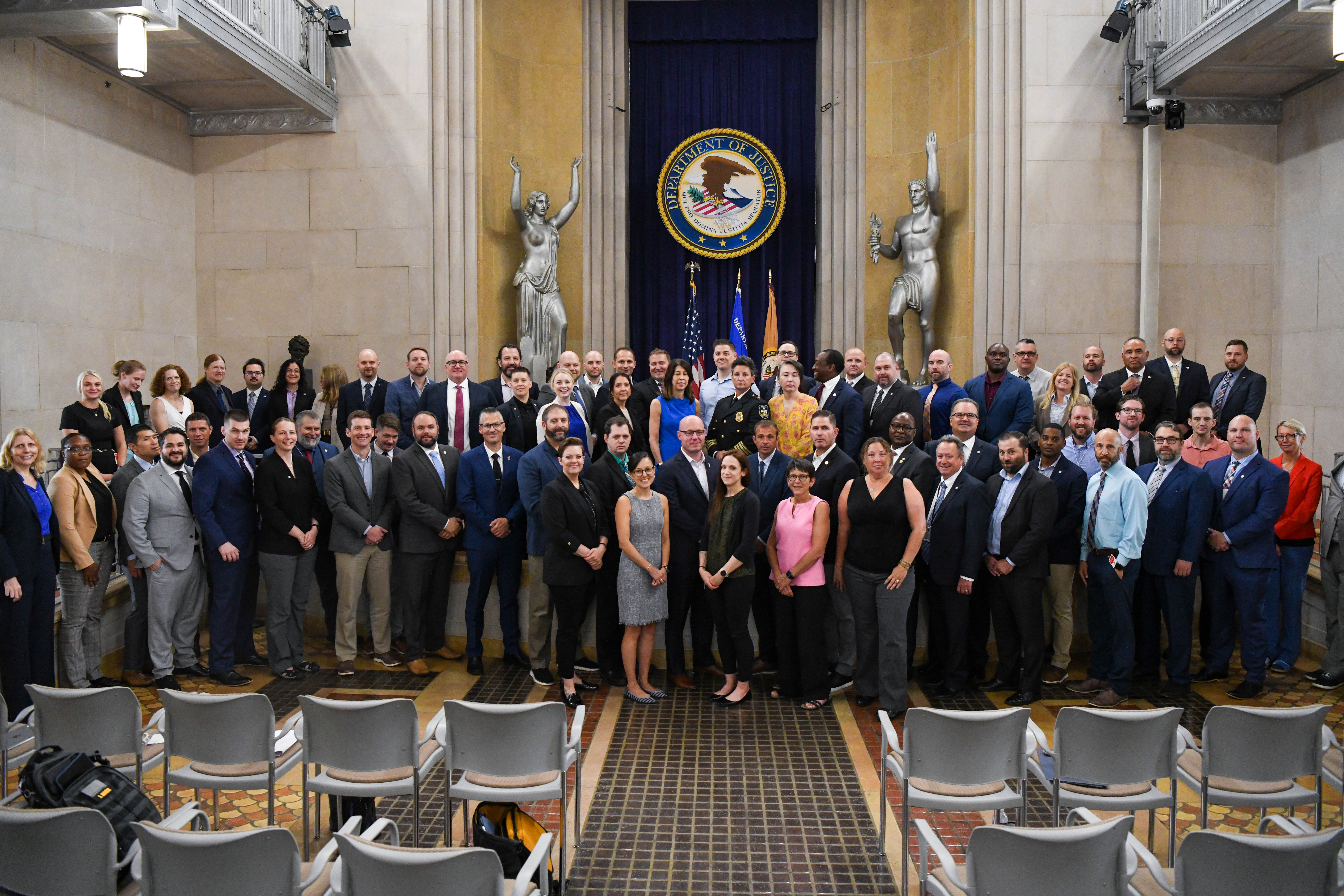 A large group of diverse professionals posed for a photo in the U.S. Department of Justice building. They stand in front of the Department of Justice seal, flanked by classical statues. The ornate room has high ceilings and marble walls. Empty chairs are visible in the foreground. The group includes men and women of various ages and ethnicities, mostly wearing formal business attire.