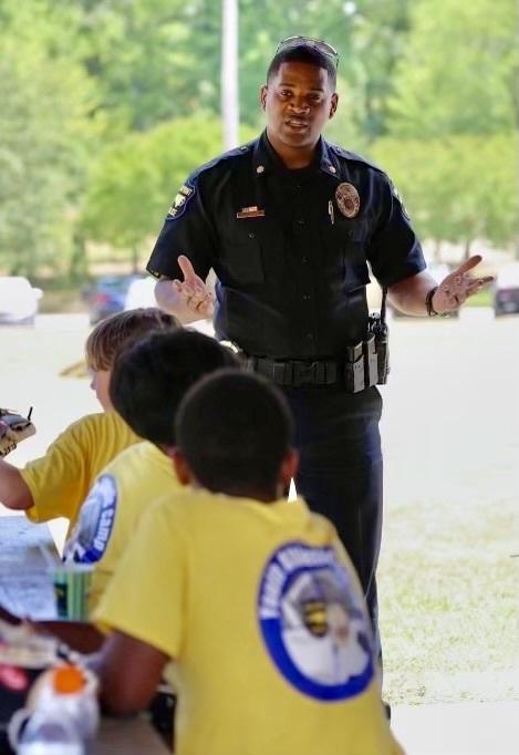 A police officer in a dark blue uniform stands gesturing with his hands while speaking to a group of children sitting on the ground. The children are wearing bright yellow shirts with a logo visible on the back. The setting appears to be outdoors, with greenery visible in the background.