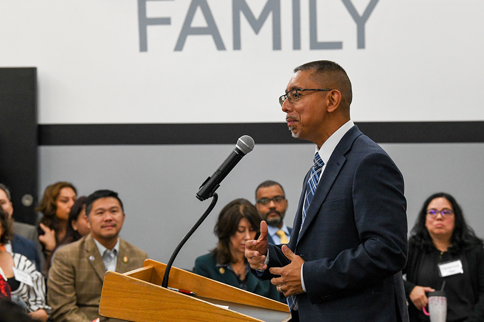A man in a navy suit speaks at a podium with "FAMILY" visible on the wall behind him. Several people are seated in the background.