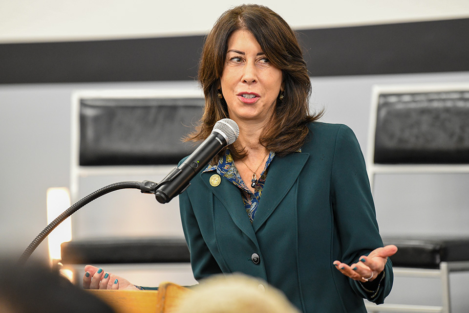 A woman with long dark hair in a teal blazer speaks into a microphone at a podium. The background shows a wall with horizontal stripes and chairs.