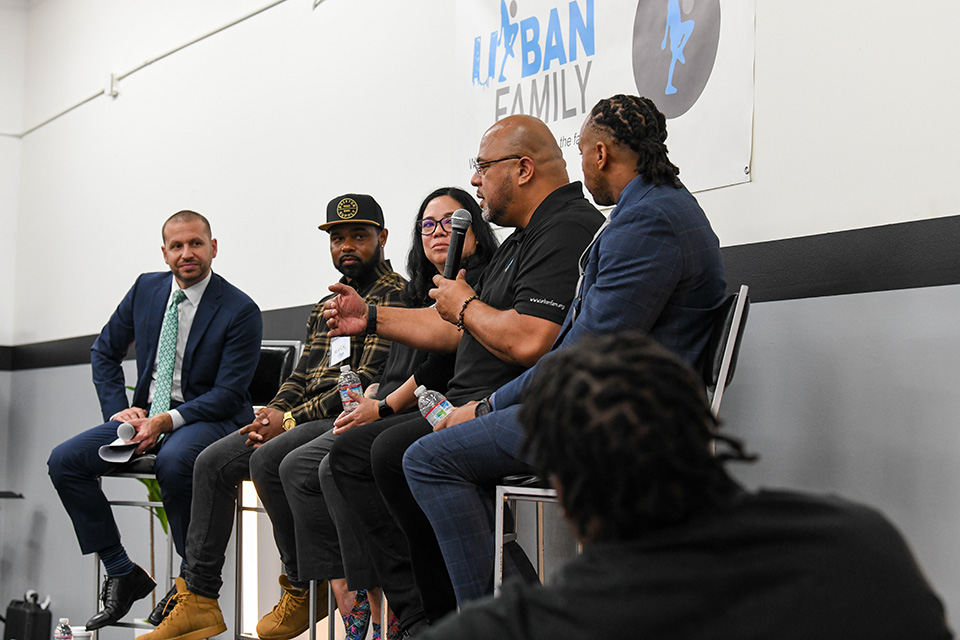 Five people seated on a raised platform, in a panel discussion. The Urban Family logo is visible on the wall behind them. The panelists include both men and women of diverse backgrounds.