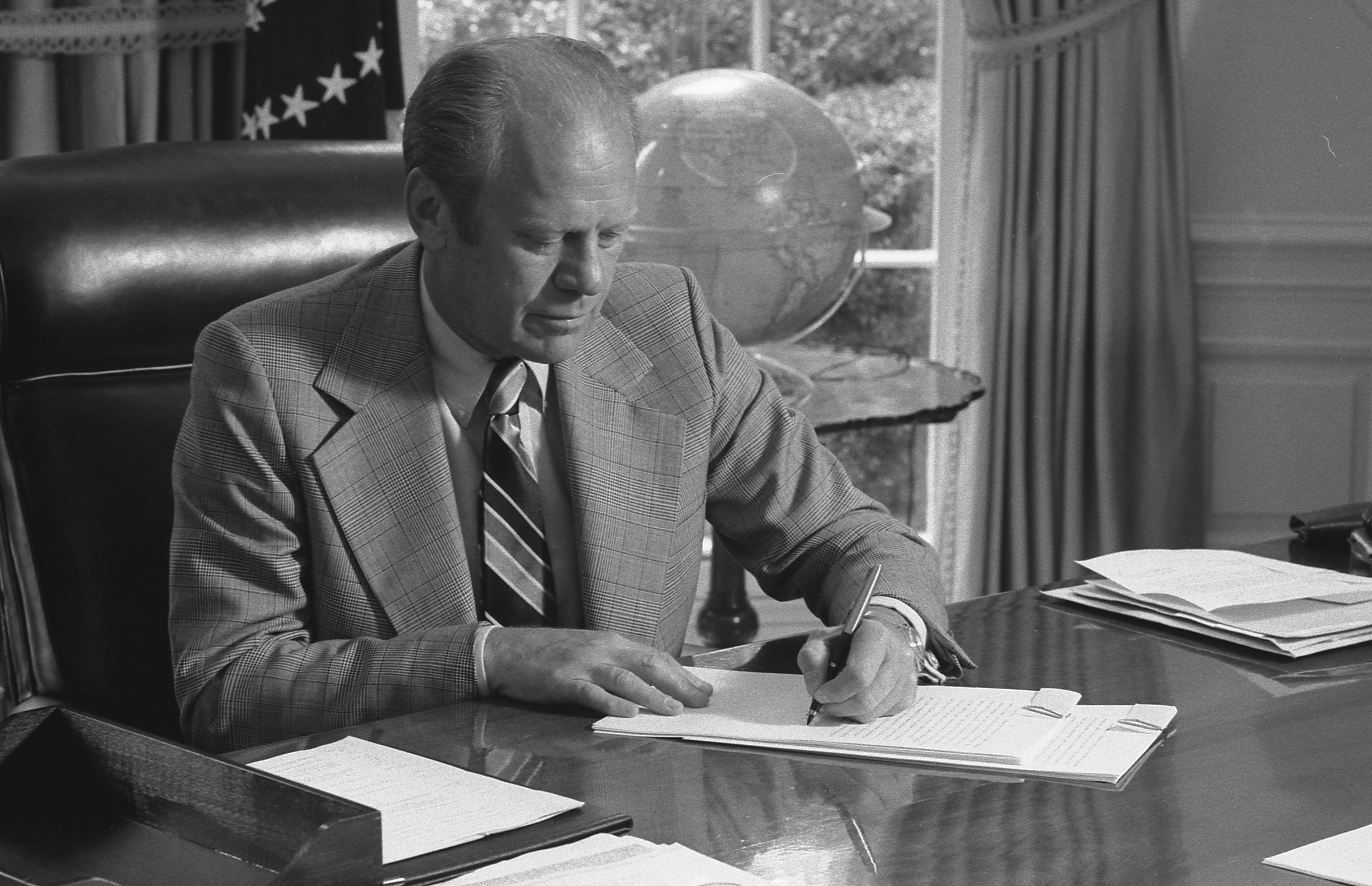 A black and white photograph shows President Gerald Ford sitting at the Resolute Desk in the Oval Office. He's wearing a plaid suit and striped tie, intently writing on papers. The American flag is visible in the background, and a globe sits on a nearby table. Ford's focused posture captures him in a moment of presidential duties, likely during his 1974-1977 term.