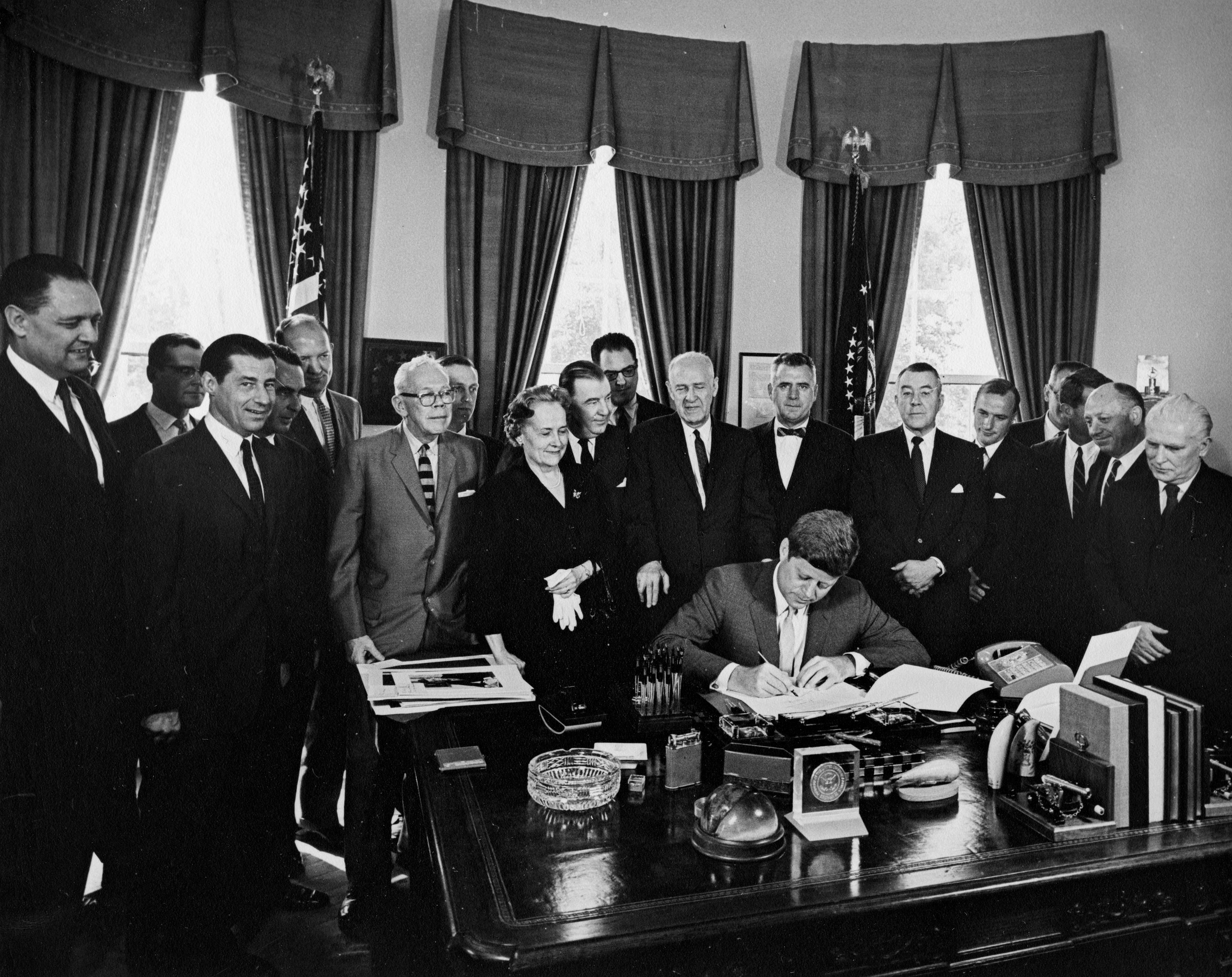 A black and white photograph shows President John F. Kennedy seated at his desk in the Oval Office, signing the Juvenile Delinquency and Youth Offenses Control Act (S. 279). He is surrounded by a group of approximately 15 men and one woman, likely lawmakers and officials, watching the signing. The desk is crowded with office items including pens, a telephone, and papers. American flags are visible in the background, framed by the distinctive curtains of the Oval Office.