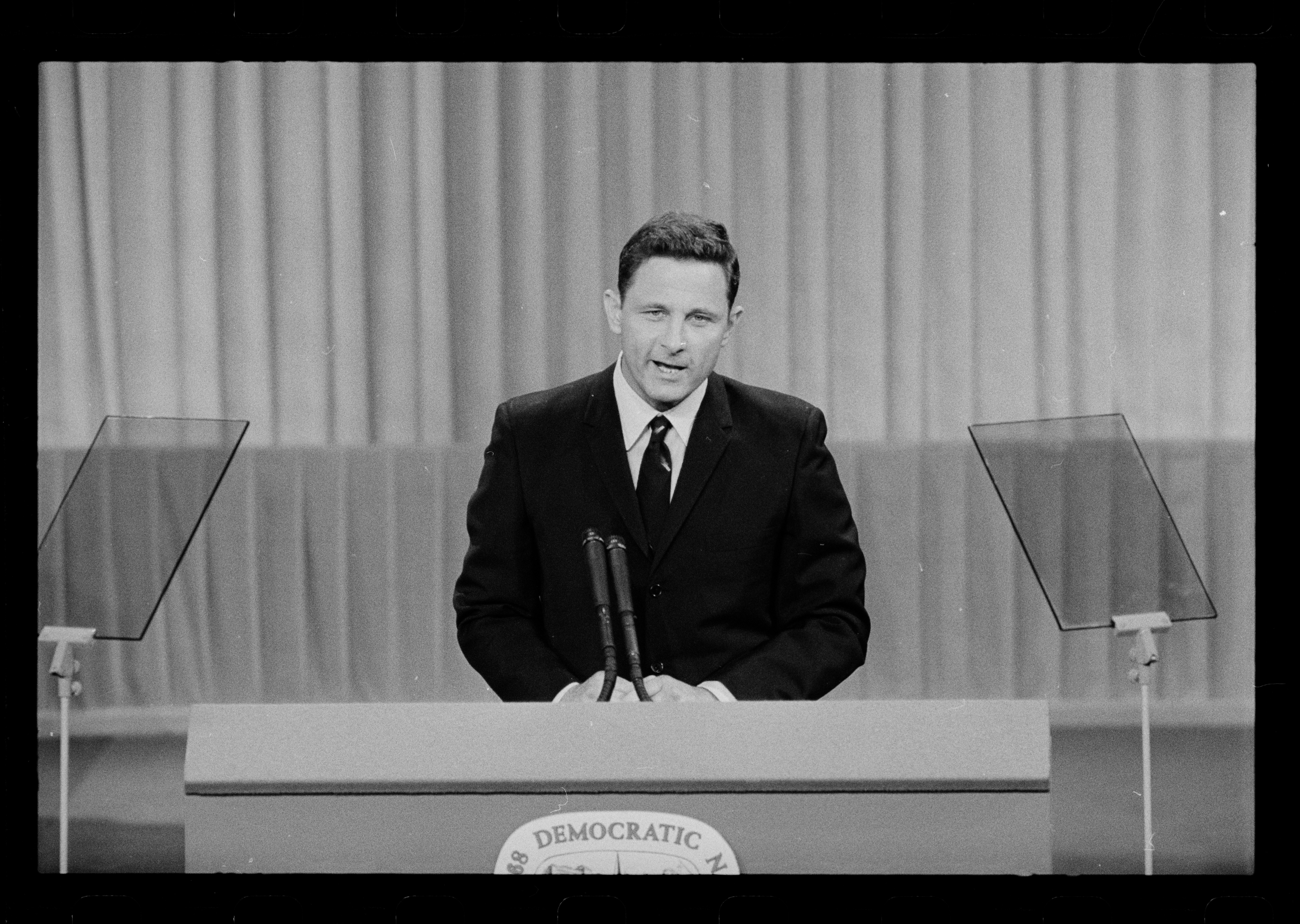 A black and white photograph shows Senator Birch Bayh in a dark suit and tie speaking at a podium. The podium has "Democratic" visible on its front, suggesting it's at a Democratic National Convention. Two transparent lectern screens flank the speaker. The man appears to be mid-speech, with his mouth open. The background is a plain curtain, typical of a stage or convention setting.