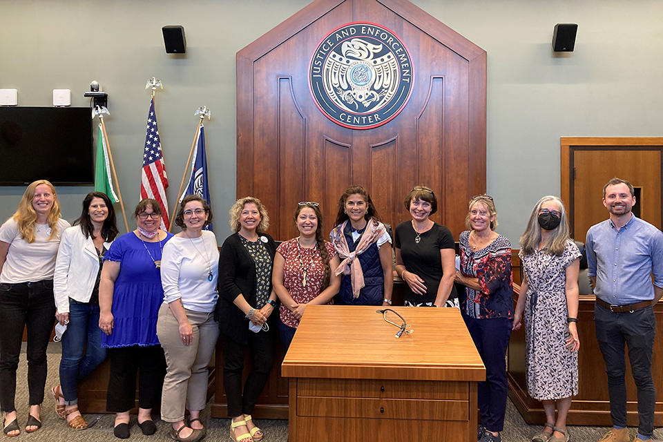 A group of eleven people standing in a room with a wooden podium and a large emblem on the wall behind them that reads 'Justice and Enforcement Center.' The group is positioned in front of three flags: one green, one American, and one blue. The room has a formal setting with a wooden door and a television screen on the left side.