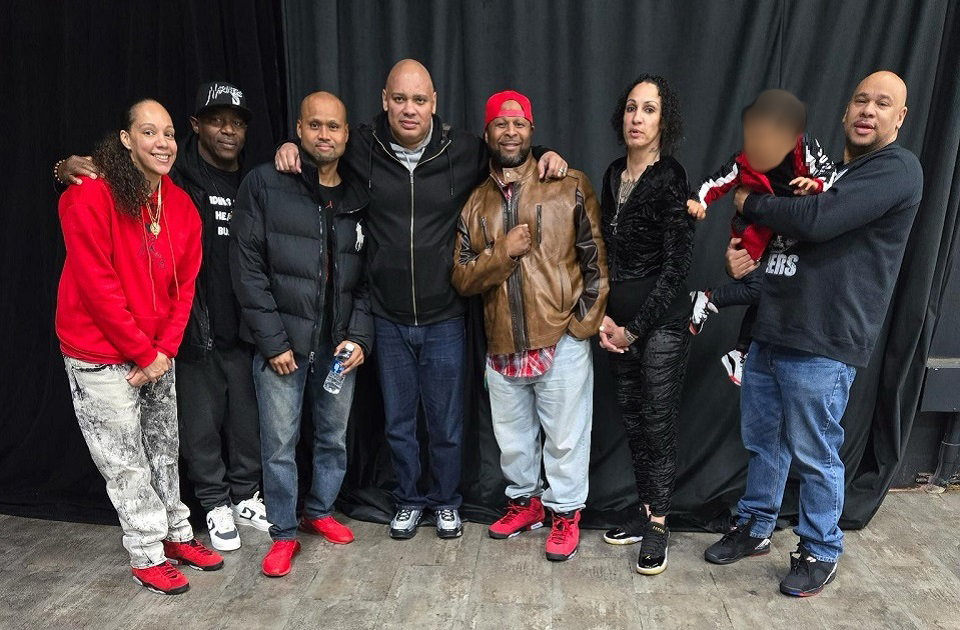 Seven people standing together in front of a black curtain. They're dressed in various outfits, including a red hoodie, a black puffer jacket, a brown leather jacket, and a black patterned outfit. One person is holding a child dressed in red and black. The group appears to be posing for a casual photo, with some wearing sneakers and others wearing casual shoes. The floor is wooden.