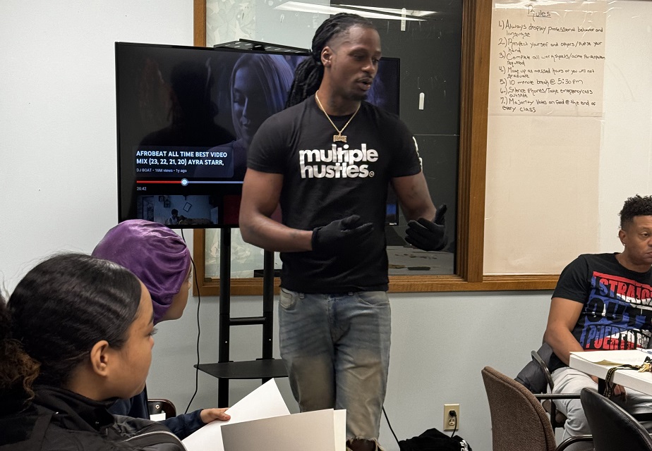 A man wearing a 'multiple hustles' t-shirt speaks to a small audience in a classroom setting. A woman in the foreground holds papers, while a TV screen and whiteboard are visible in the background, suggesting an educational or training environment.
