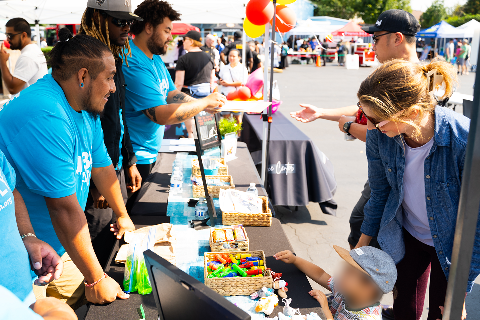 Volunteers in bright blue shirts engage with attendees at a lively street fair booth. The table displays colorful items and informational materials, while balloons and tents are visible in the background, creating a festive atmosphere.