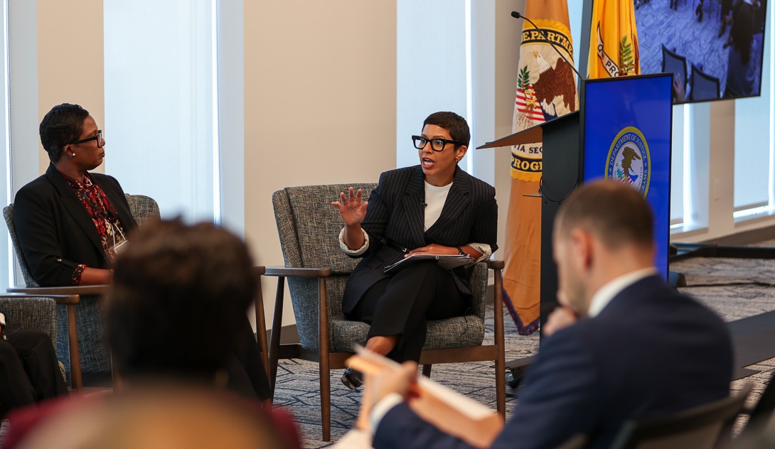 "A panel discussion with speakers seated in gray armchairs, with Department of Justice flags and seal visible in the background. One speaker in a black blazer and white top is gesturing while speaking
