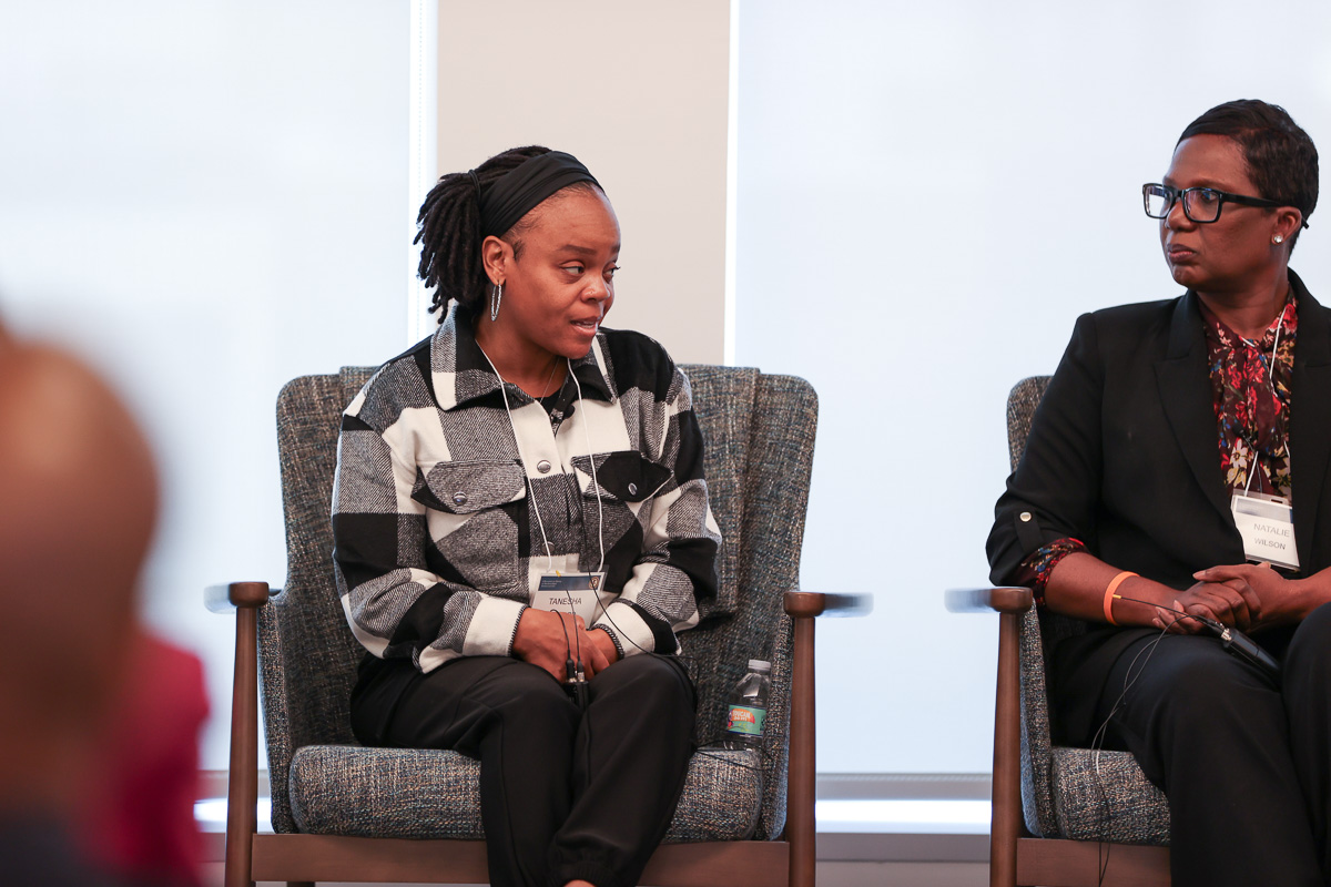 Two participants in a panel discussion seated in gray armchairs. One person wears a black and white checkered jacket and has dreadlocks, while another wears a black blazer with a patterned blouse