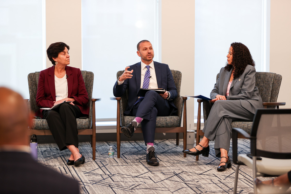 Three speakers seated in gray armchairs during a panel discussion. From left to right: a person in a burgundy blazer, a person in a navy suit with a striped tie, and a person in a gray suit