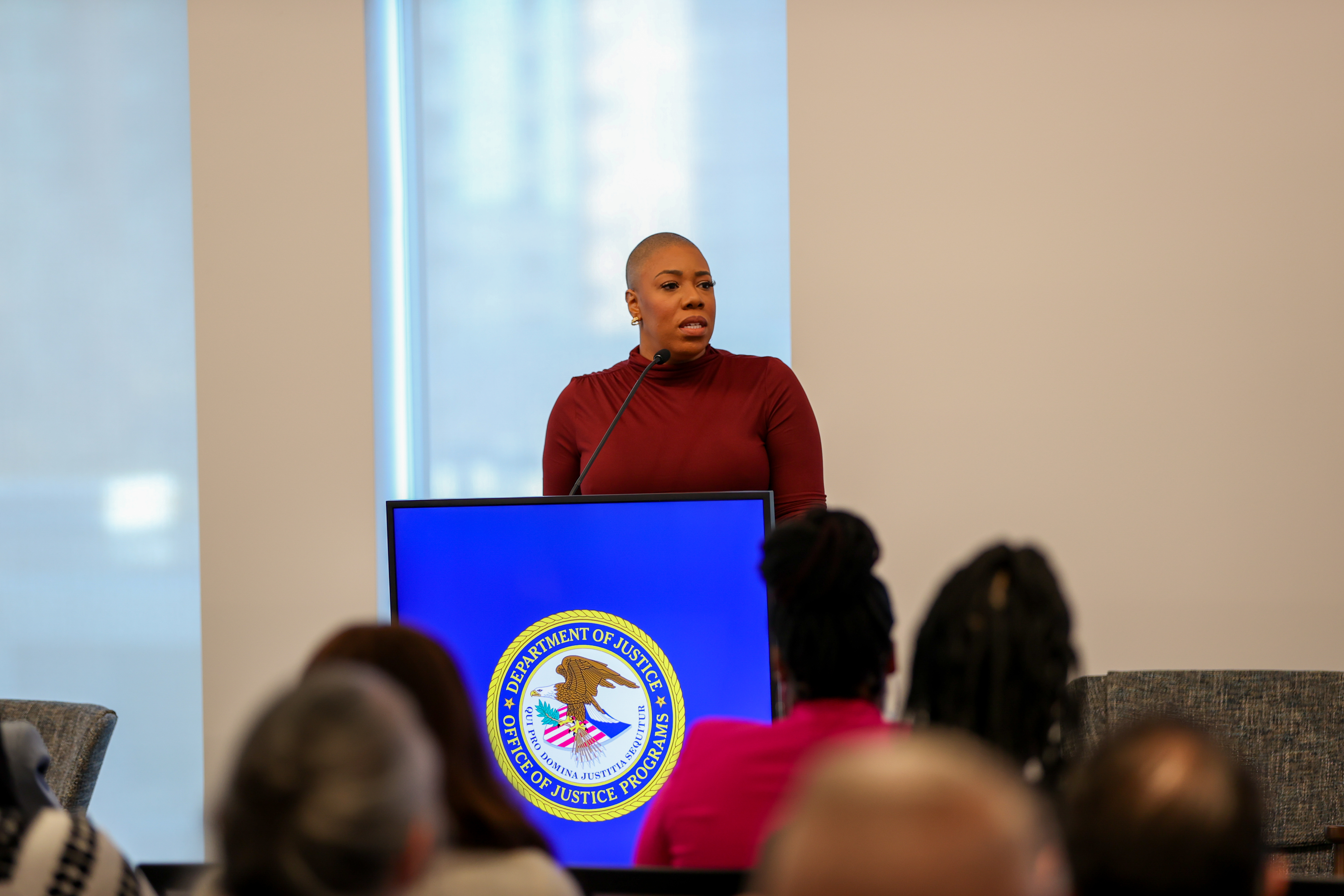 A person in a burgundy turtleneck speaking at a podium with the Department of Justice Office of Justice Programs seal displayed on a blue screen behind them