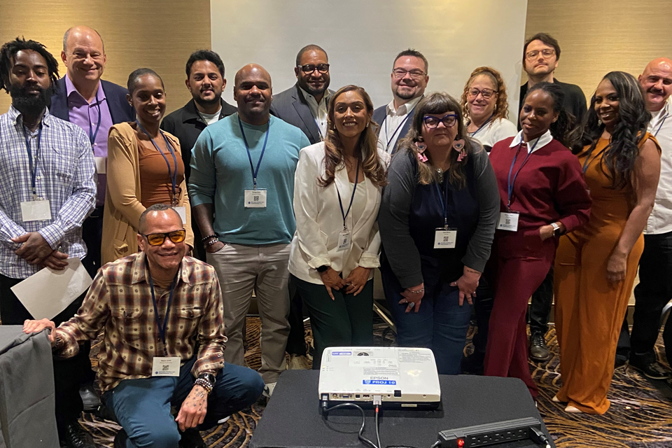 A group photo of approximately fourteen people from the National Reentry Housing Incubator program. The group is arranged in front of a neutral wall, with a projector visible on a table in the foreground. Attendees are dressed in professional attire ranging from business casual to formal, including outfits in colors like purple, turquoise, white, burgundy, and orange.