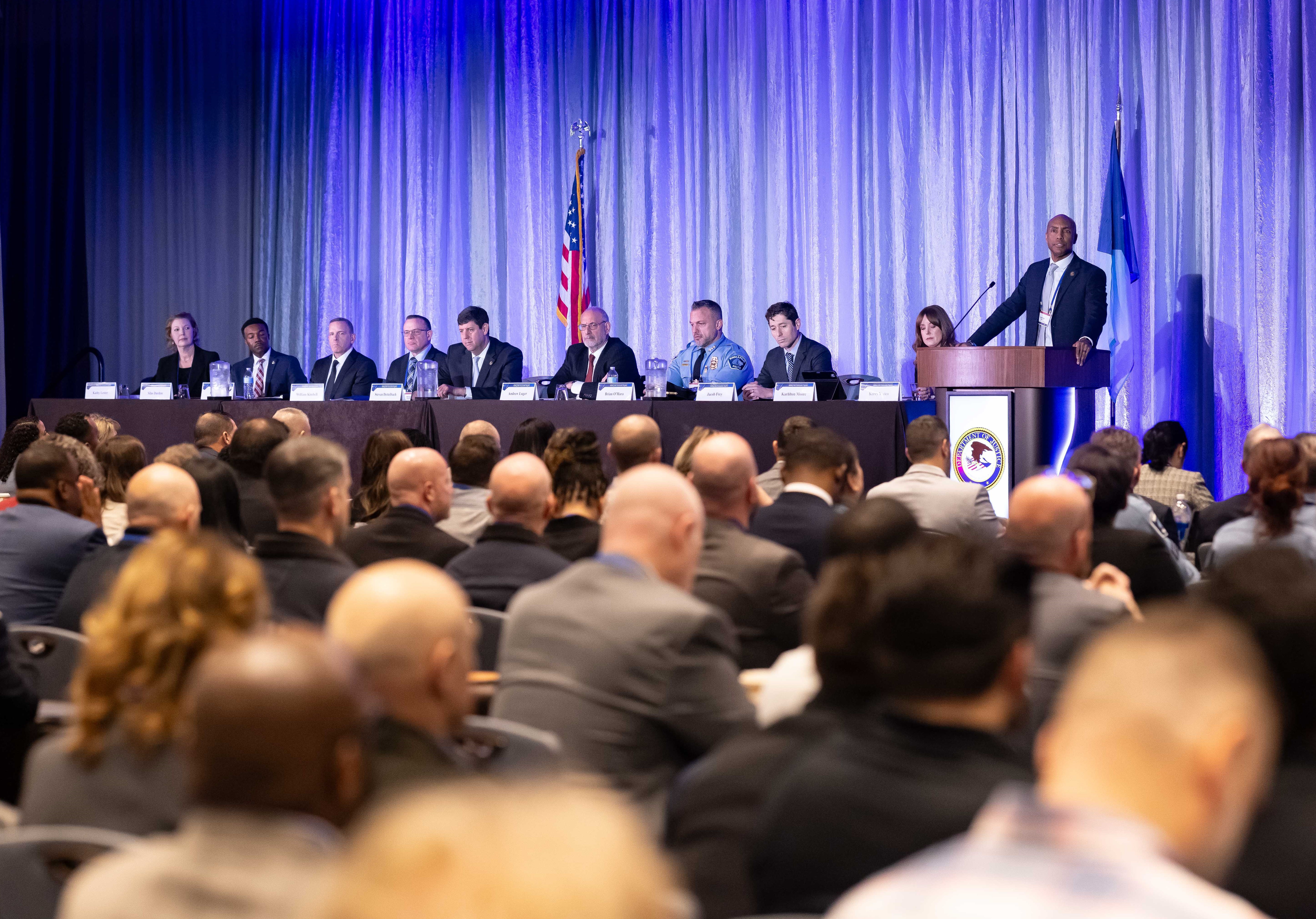 A panel discussion at the National Public Safety Partnership (PSP) Summit, showing multiple law enforcement and justice officials seated at a long table on stage with the American flag displayed. A speaker stands at a podium while a full audience of public safety professionals watches.