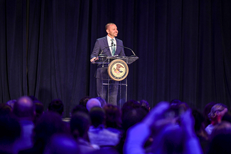 A man in a navy suit and striped tie speaking at a clear podium with the Department of Justice seal, with an audience visible in the foreground.