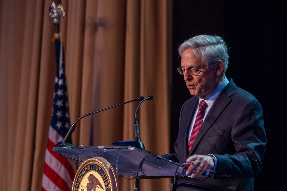 Attorney General Garland in a dark suit and red tie speaking at a clear podium with the Department of Justice seal and American flag visible.