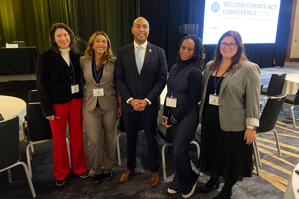 Five staff members from Impact Justice and the Bureau of Justice Assistance pose in front of a Second Chance Act Conference banner. They are wearing professional attire including blazers, suits, and professional dresses in colors including black, red, gray, and navy.