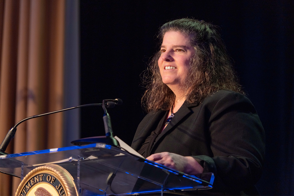 A woman with long dark curly hair wearing a black blazer speaking at a podium with the Department of Justice seal visible.