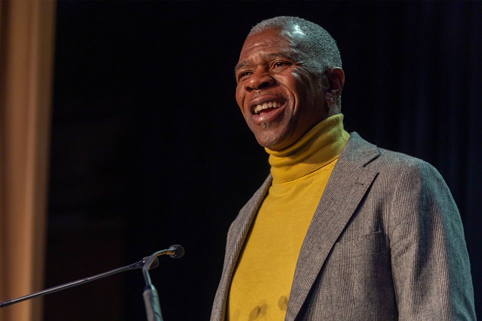 A man wearing a yellow turtleneck and gray blazer smiling while speaking at a podium.