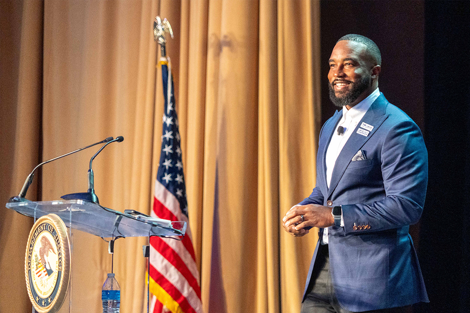 A man wearing a navy suit and white shirt smiling while standing at a podium with the Department of Justice seal and American flag visible.