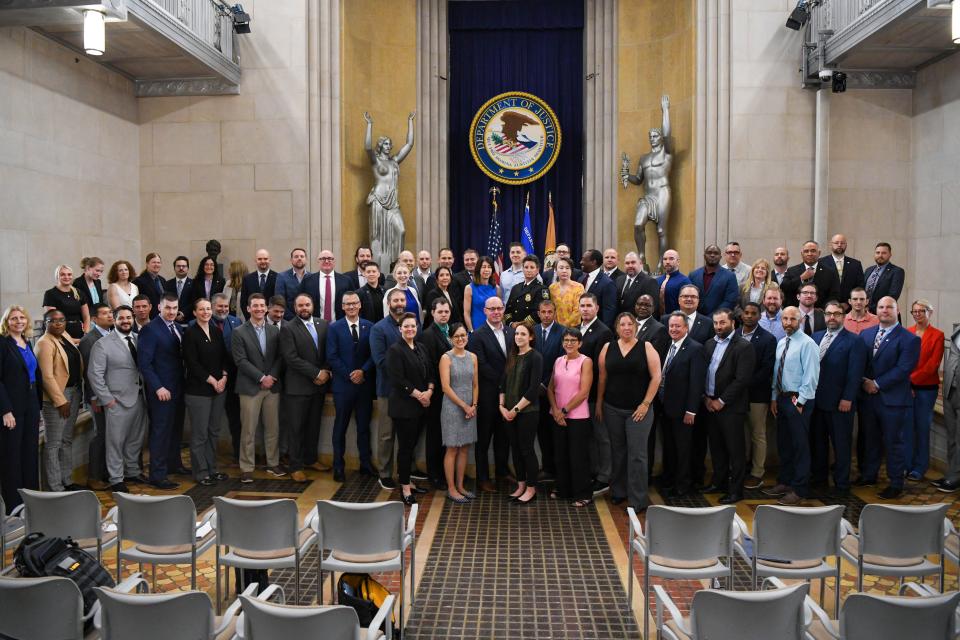 A large group of diverse professionals posed for a photo in the U.S. Department of Justice building. They stand in front of the Department of Justice seal, flanked by classical statues. The ornate room has high ceilings and marble walls. Empty chairs are visible in the foreground. The group includes men and women of various ages and ethnicities, mostly wearing formal business attire.