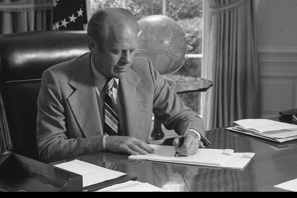 A black and white photograph shows President Gerald Ford sitting at the Resolute Desk in the Oval Office. He's wearing a plaid suit and striped tie, intently writing on papers. The American flag is visible in the background, and a globe sits on a nearby table. Ford's focused posture captures him in a moment of presidential duties, likely during his 1974-1977 term.