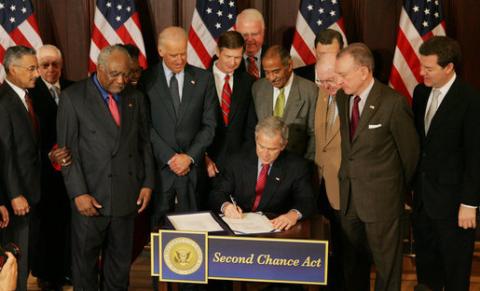 President Bush seated at desk signs Second Chance Act. Standing Congressional representatives look on. US flags in background