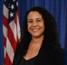 Professional government portrait of a woman with long, curly dark hair wearing a black blazer with a v-neck, standing in front of a blue curtain backdrop with an American flag. She is smiling warmly at the camera and wearing a delicate gold necklace.